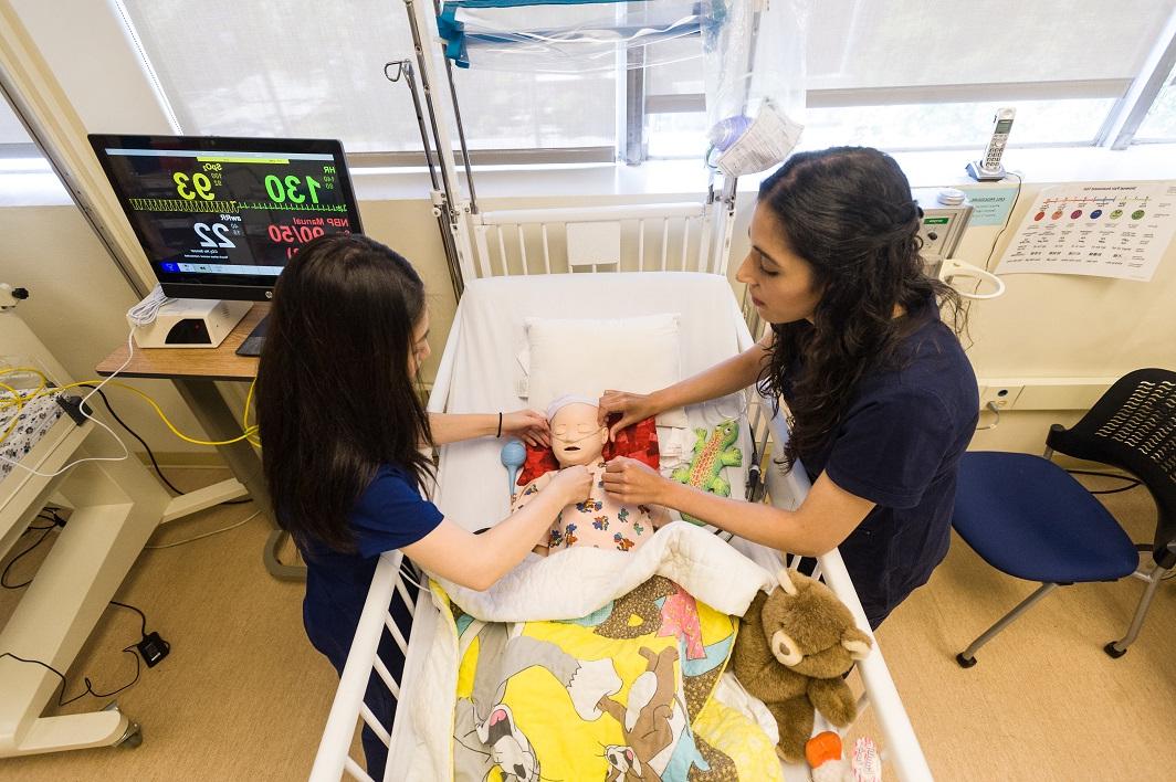 Two nursing students in scrubs practice skills on a baby doll in a crib.